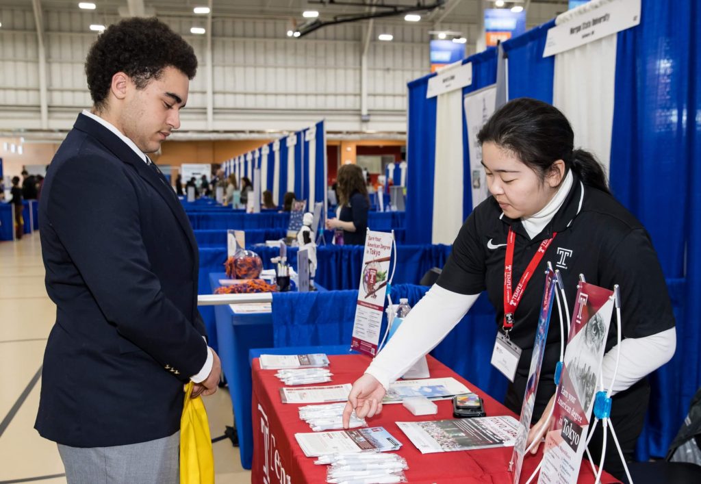 An R-MA student attentively listens as a college representative points to materials on a table, displaying options for studying abroad