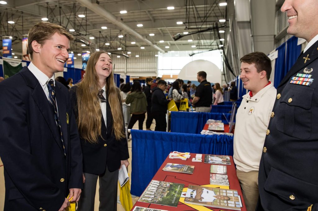 R-MA students chat and laugh with college representatives in uniform at a college fair, with various booths in the background