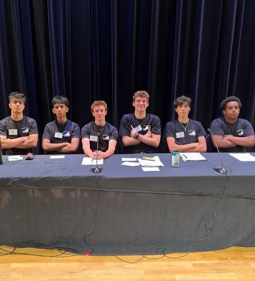 Six students sit at a long table with microphones, arms crossed, looking confident