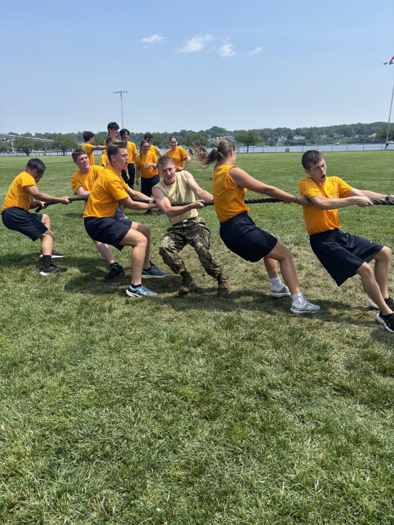 Students in yellow shirts compete in a tug-of-war, straining hard together on grass field
