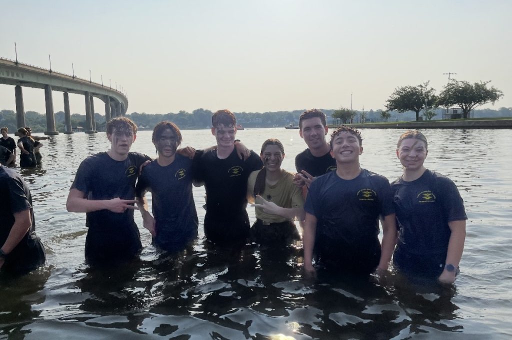 Wet and muddy students stand in river, smiling with camaraderie, faces painted, arms around teammates