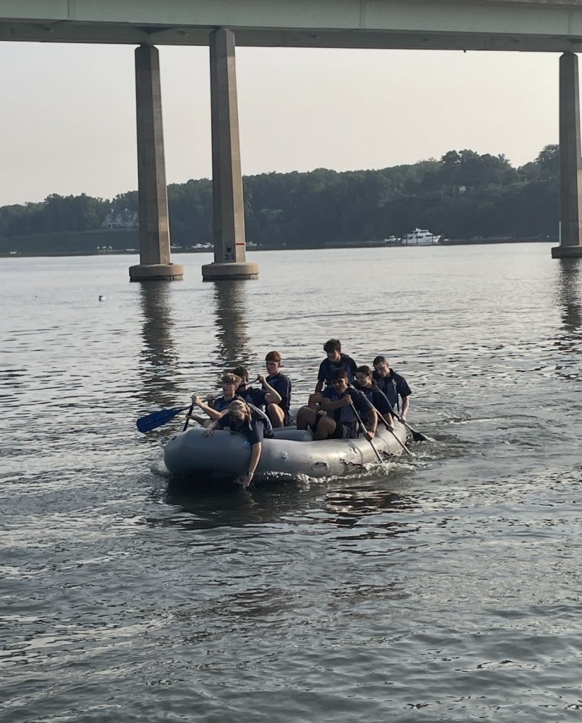 A team paddles an inflatable raft under a bridge, working together across calm river water