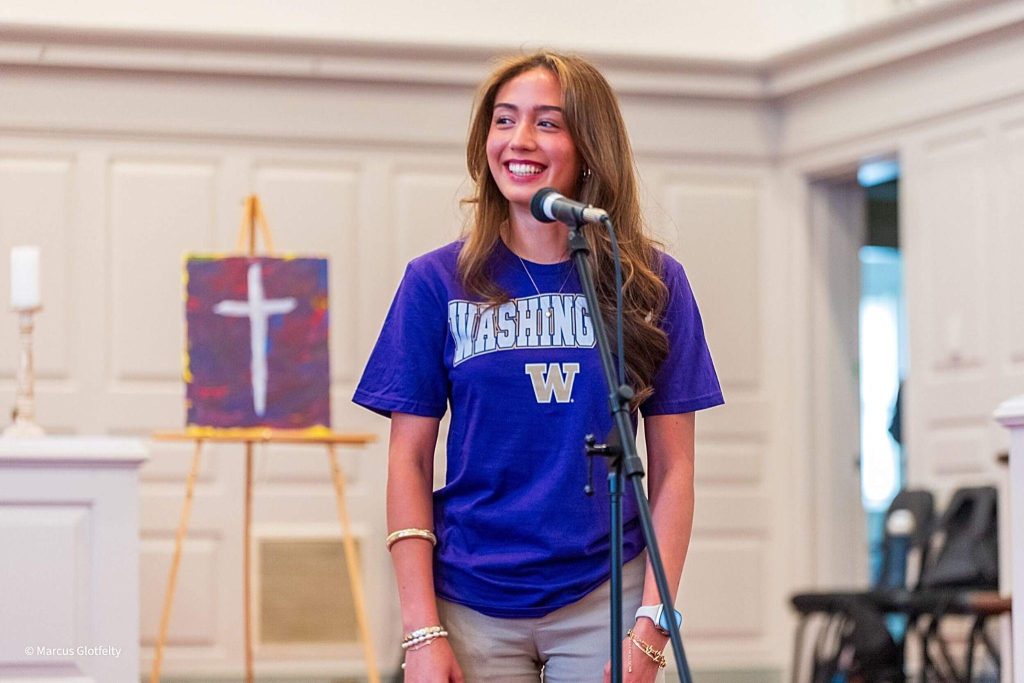 An R-MA student wearing a college shirt while speaking at a microphone during the R-MA college T-shirt reveal event.