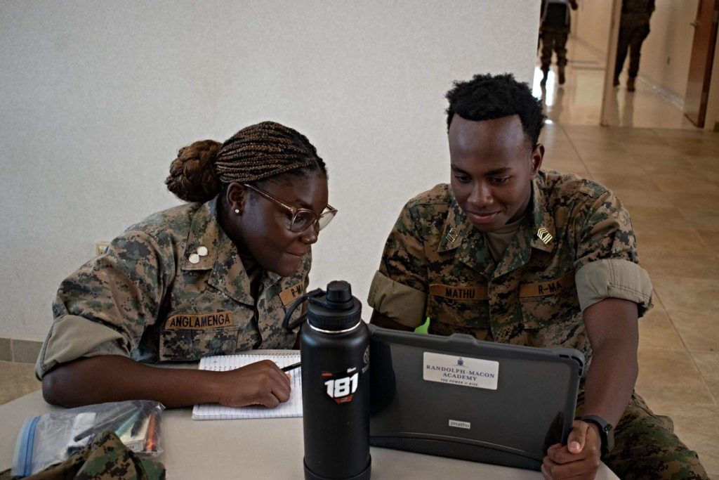 A female R-MA cadet taking notes while working with a classmate on a laptop during an academic activity.