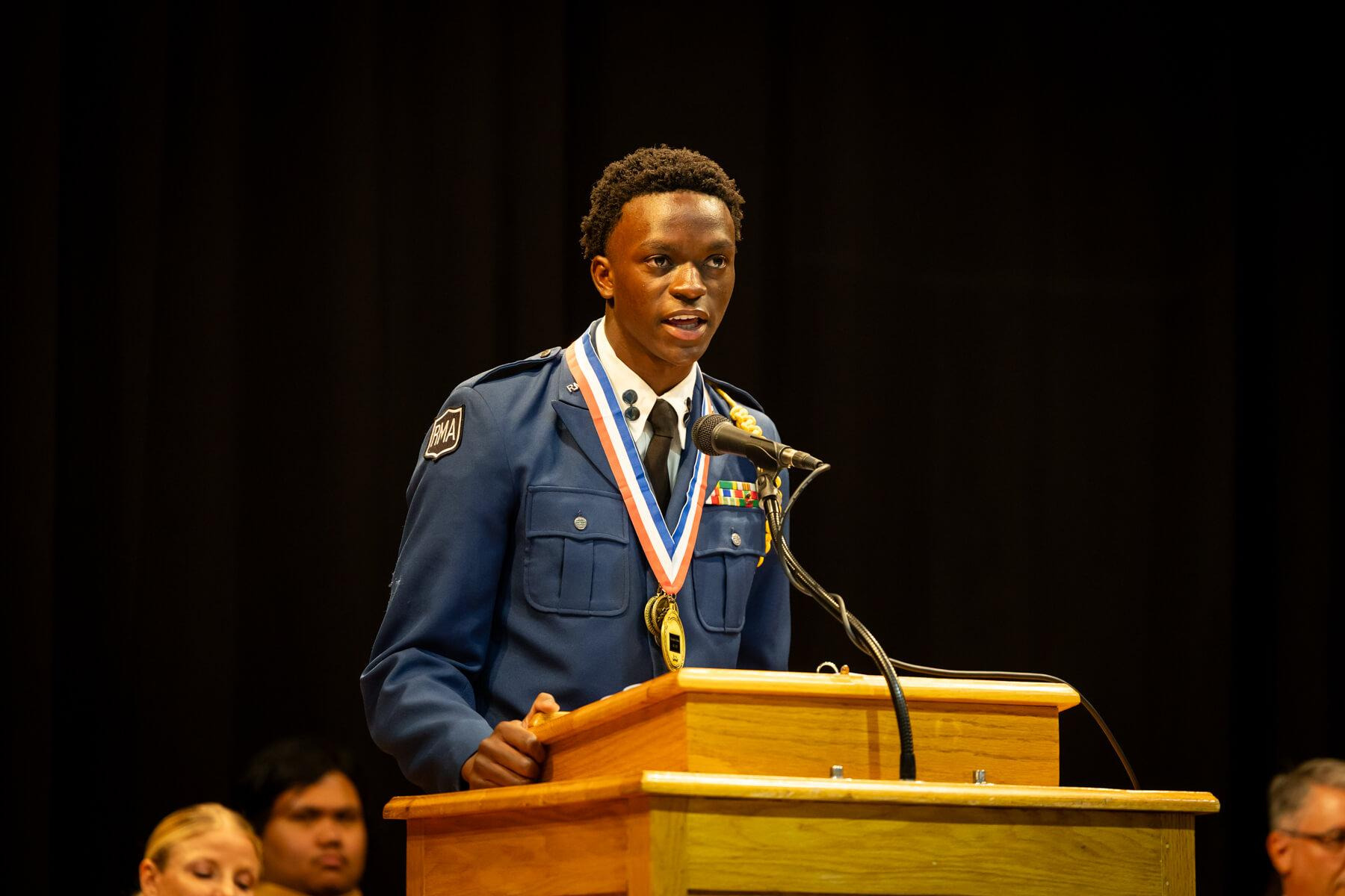 R-MA cadet speaking at a podium during a school ceremony, demonstrating confident communication.